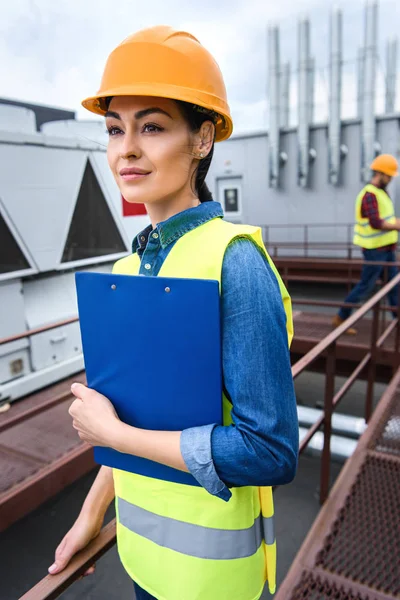 Architektin im Helm hält Klemmbrett auf Dach, männlicher Kollege dahinter — Stockfoto