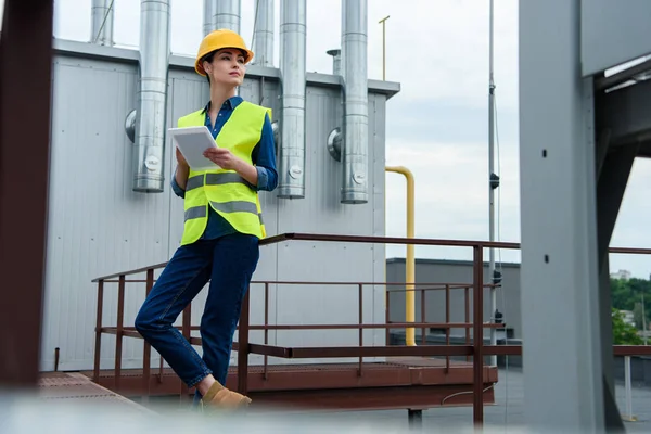 Professional female engineer in safety vest and helmet using tablet on industrial construction — Stock Photo