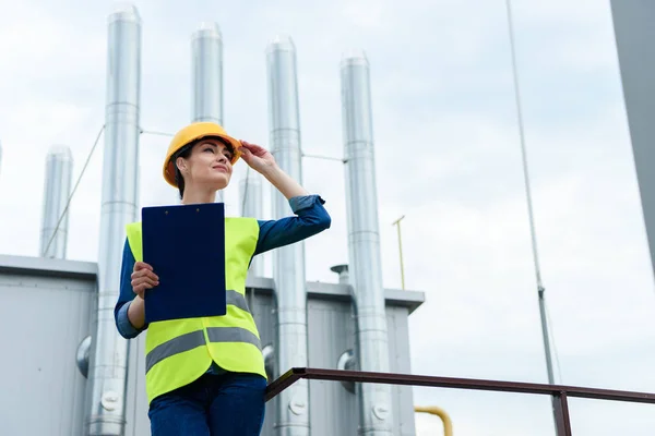 Ingénieur attrayant en gilet de sécurité et casque écriture dans le presse-papiers sur la construction industrielle — Photo de stock