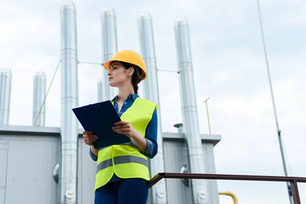 Beautiful female builder in safety vest and hardhat writing in clipboard on industrial construction — Stock Photo