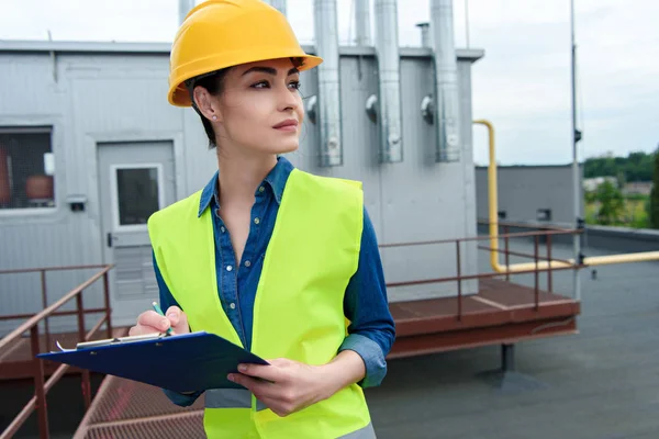 Hermosa mujer ingeniera escribiendo en portapapeles en la construcción industrial - foto de stock