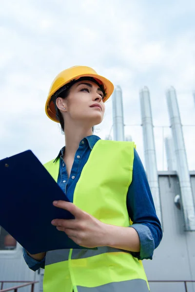 Vue du bas de belle architecte féminine en gilet de sécurité et écriture de casque dans le presse-papiers — Photo de stock