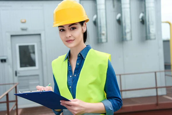 Attractive engineer in safety vest and hardhat writing in clipboard — Stock Photo