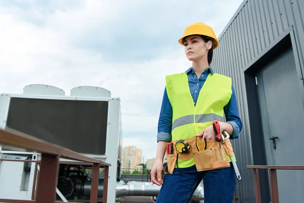 Ingeniera femenina con cinturón de herramientas posando en chaleco de seguridad y sombrero de fuerza - foto de stock