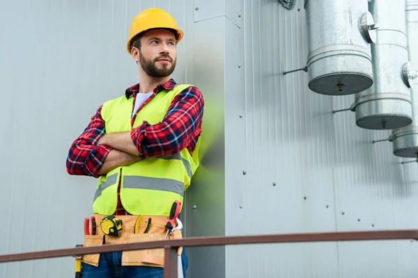 Männlicher Berufsingenieur mit Werkzeuggurt posiert mit verschränkten Armen an der Wand — Stockfoto