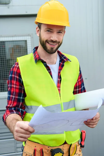 Ingeniero sonriente en chaleco de seguridad y casco trabajando con planos - foto de stock