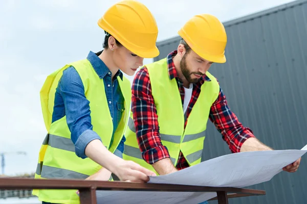 Professional architects in safety vests and helmets working with blueprints on roof — Stock Photo