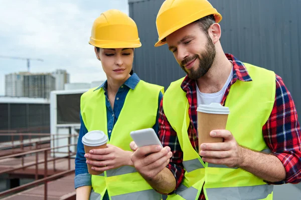 Engineers in helmets using smartphone on coffee break on roof — Stock Photo