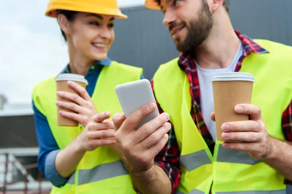 Architects in safety vests and helmets using smartphone on coffee break — Stock Photo