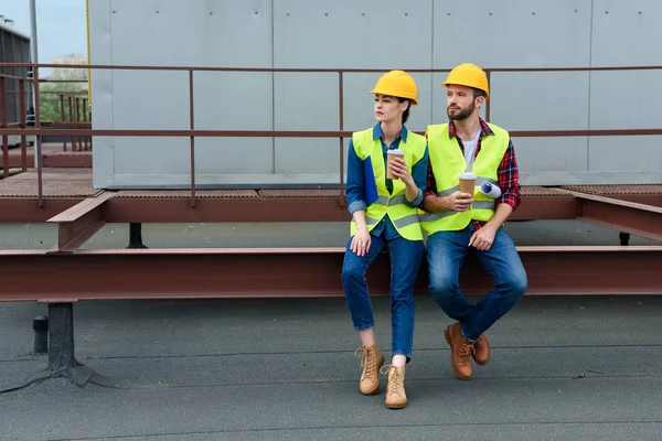 Arquitectos masculinos y femeninos en sombreros con plano en la pausa de café sentado en el techo - foto de stock