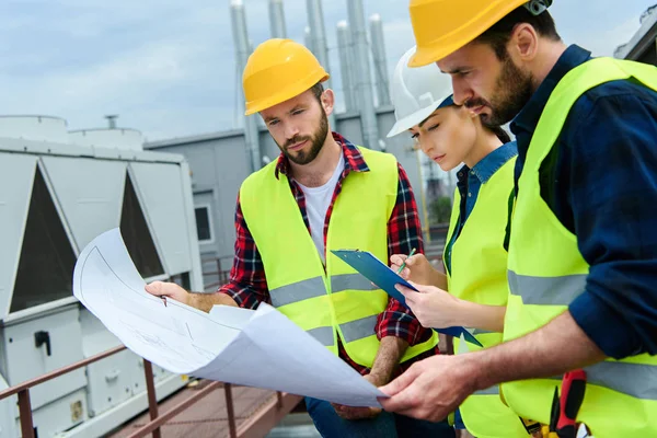 Professional engineers in safety vests and hardhats working with blueprints and clipboard on roof — Stock Photo