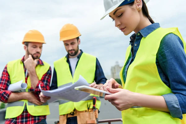 Professional engineers in safety vests and helmets working with digital tablet and blueprints — Stock Photo