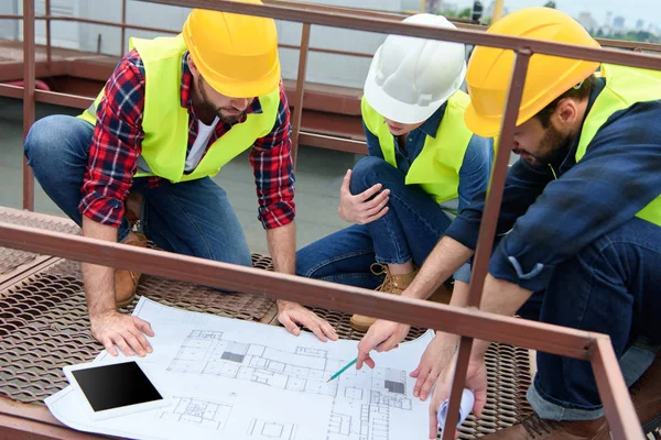 Três engenheiros em coletes de segurança e chapéus duros trabalhando com plantas no telhado — Fotografia de Stock
