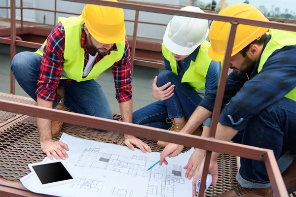 Tres ingenieros en chalecos de seguridad y sombreros de trabajo con planos en el techo - foto de stock