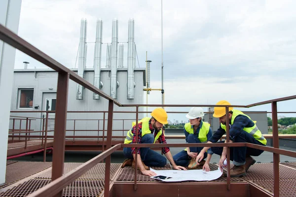 Three engineers in helmets working with blueprints on roof together — Stock Photo