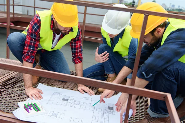 Drei Architekten in Harthüten arbeiten mit Bauplänen auf dem Dach — Stockfoto