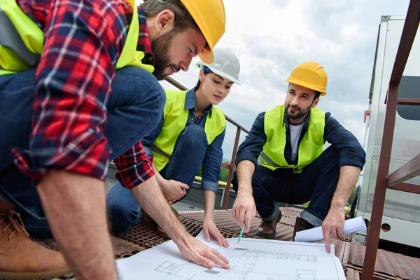 Three engineers in hardhats working with blueprints on roof — Stock Photo