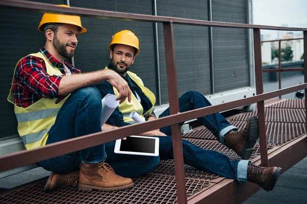 Архітектори в хардхаусах з кресленнями та цифровим планшетом, що сидить на будівництві — стокове фото