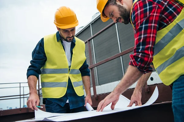 Arquitectos en chalecos de seguridad y sombreros de trabajo con planos en el techo - foto de stock