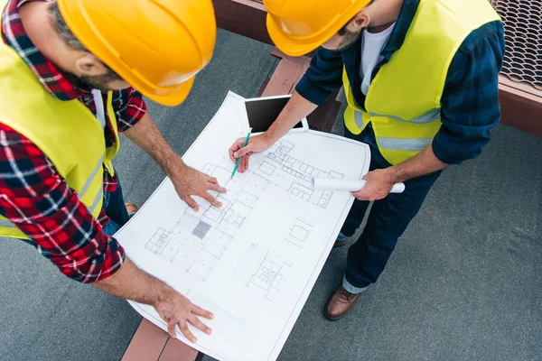 Vista aérea de los arquitectos en chalecos de seguridad y cascos dibujando sobre planos - foto de stock