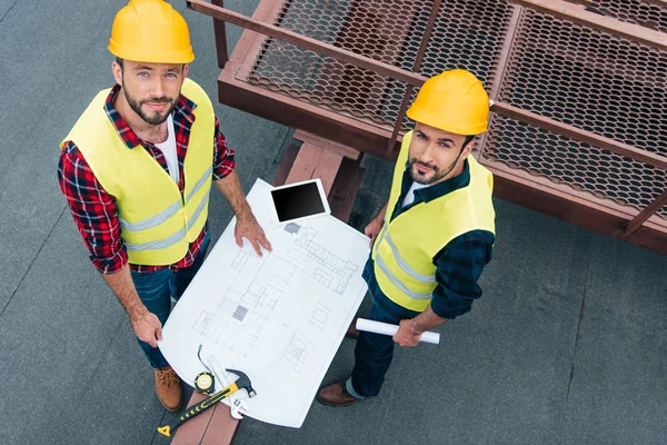 Vista aerea di ingegneri maschi in giubbotti e caschi di sicurezza che lavorano con cianografie, tablet digitali e strumenti sul tetto — Foto stock