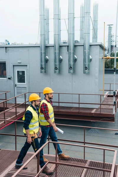 Construct coworkers in safety vests and helmets with blueprint and tool belt on roof — Stock Photo