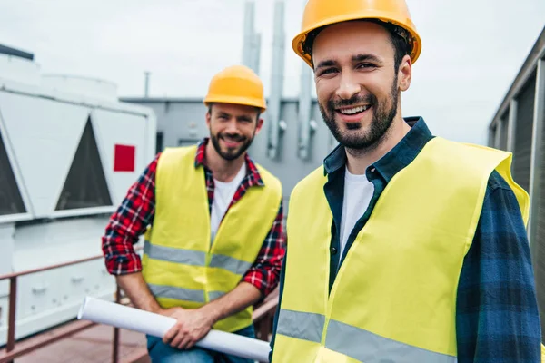 Smiling engineers in safety vests and helmets with blueprint on roof — Stock Photo