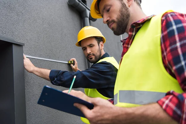 Male engineers in safety vests and helmets working with measuring tape and clipboard near wall — Stock Photo