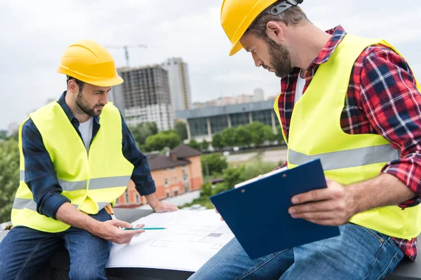 Dos ingenieros en cascos que trabajan con planos y portapapeles en el techo - foto de stock