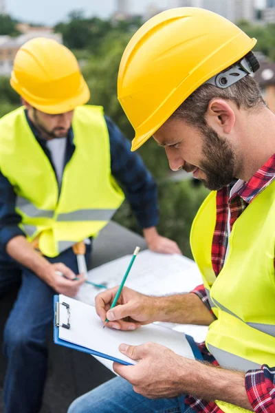 Arquitectos en chalecos de seguridad y sombreros, uno de ellos escrito en el portapapeles - foto de stock