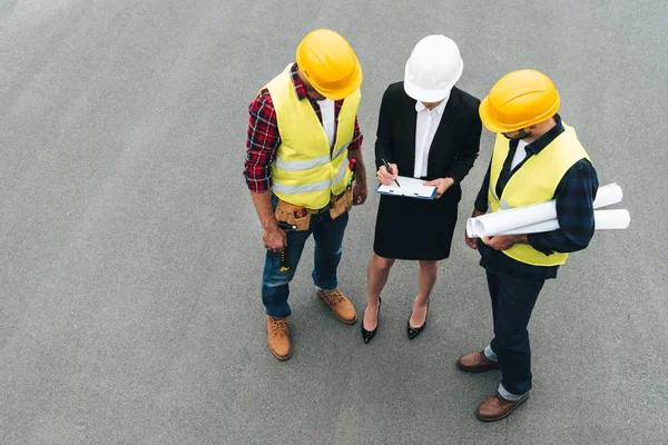 Vista aérea de la arquitecta y los trabajadores masculinos en sombreros de trabajo con portapapeles y planos - foto de stock