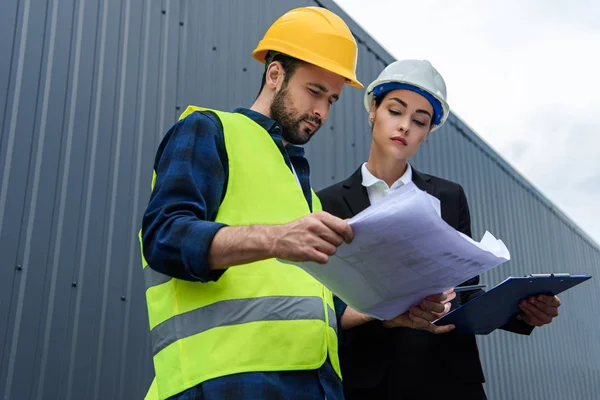 Mujer ingeniera y trabajadora masculina en cascos con planos en construcción — Stock Photo