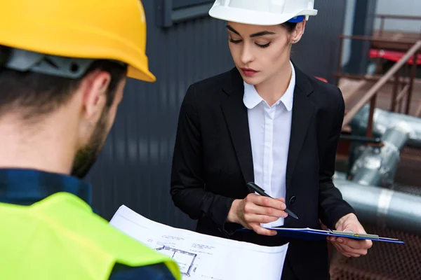 Enfoque selectivo de la arquitecta y el trabajador masculino en cascos con plano y portapapeles en la construcción - foto de stock