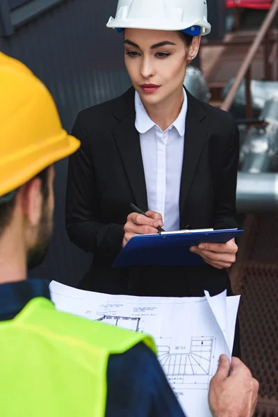 Selective focus of architect and worker in helmets with blueprint and clipboard on construction — Stock Photo