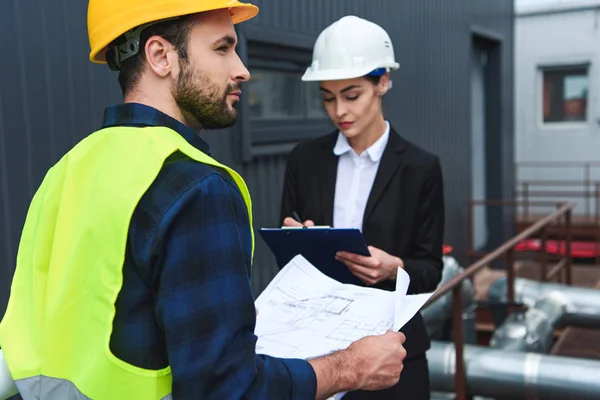 Selective focus of architect and builder in helmets with blueprint and clipboard on construction — Stock Photo