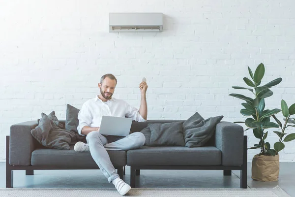 Mann schaltet Klimaanlage mit Fernbedienung ein, während er Laptop auf Sofa benutzt — Stockfoto