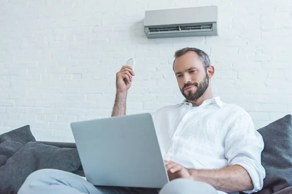 Bearded man turning on air conditioner with remote control while using laptop — Stock Photo