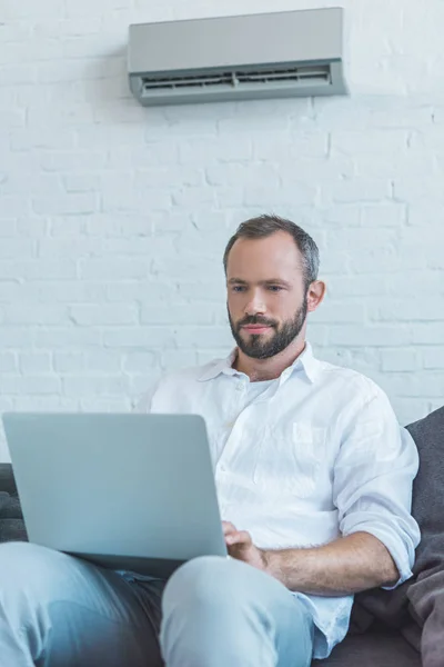 Bearded man using laptop at home, with air conditioner on wall behind — Stock Photo