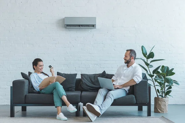Пара вмикає кондиціонер під час літньої спеки, сидячи на дивані з книгою та ноутбуком — стокове фото