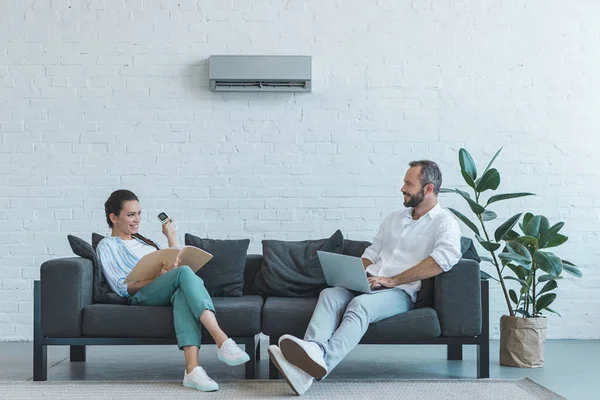 Pareja encendiendo el aire acondicionado durante el calor del verano mientras está sentado en el sofá con libro y portátil - foto de stock