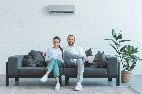 Couple sitting on sofa during the summer heat at home with ficus and air conditioner on wall — Stock Photo