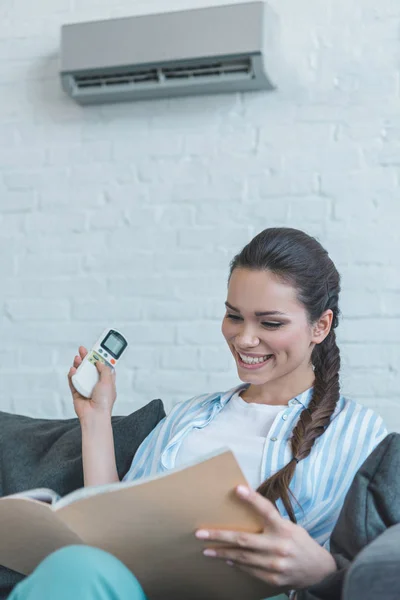 Femme souriante allumant le climatiseur avec télécommande tout en lisant le livre à la maison — Photo de stock