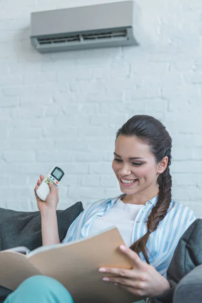 Smiling woman turning on air conditioner with remote control while reading book at home — Stock Photo