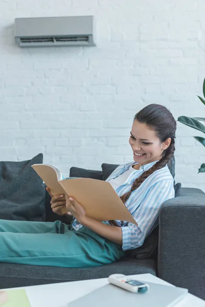 Happy woman reading book on sofa, with air conditioner on wall — Stock Photo