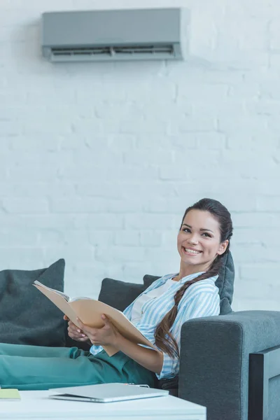 Mujer sonriente con libro sentado en el sofá, con aire acondicionado en la pared - foto de stock