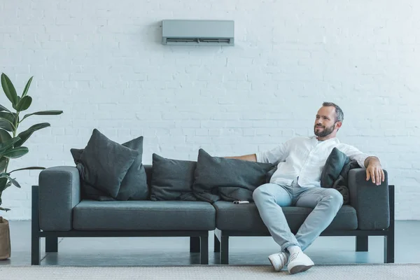 Homme assis sur canapé gris avec télécommande, climatiseur au mur — Photo de stock