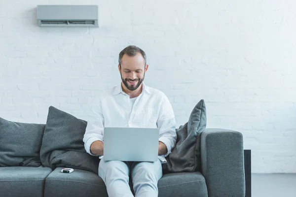 Smiling man using laptop and sitting on sofa, with air conditioner on wall — Stock Photo