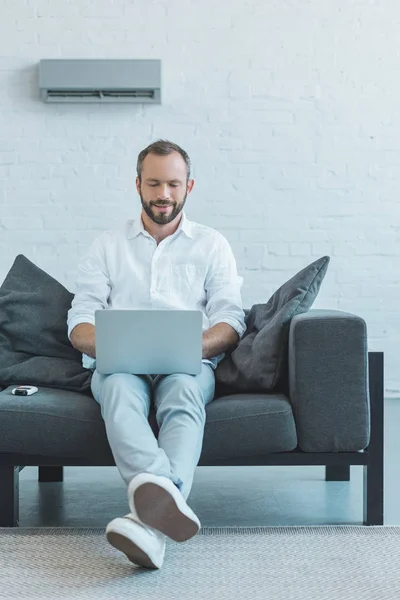 Homme barbu utilisant un ordinateur portable sur le canapé, avec climatiseur sur le mur — Photo de stock