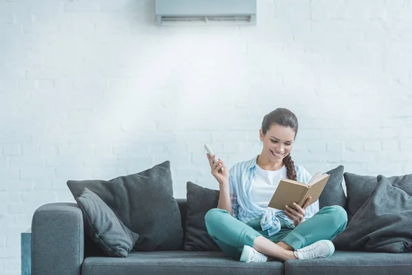 Happy woman reading book while turning on air conditioner with remote control at home — Stock Photo