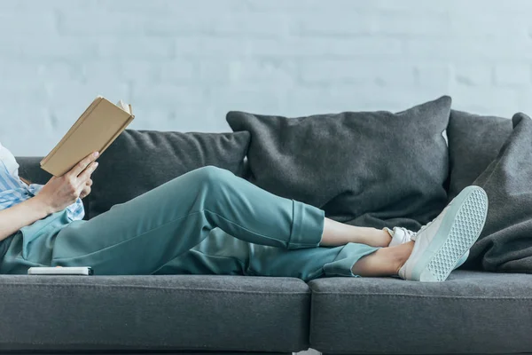 Ausgeschnittene Ansicht einer Frau, die es sich auf dem Sofa gemütlich macht und Buch liest — Stockfoto